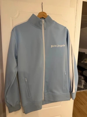 Palm angels kofta  - Snygg ljusblå hoodie från Palm Angels med vit dragkedja och vita detaljer längs ärmarna. Klassisk hög krage och två fickor med dragkedja framtill. Tillverkad i ett mjukt och stretchigt material som känns sportigt och trendigt. Palm Angels-logga i vitt på bröstet.