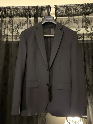 Mörkblå kavaj från J.Lindeberg - Nypris: 4200 kr Mörkblå, HELT NY kavaj från J.Lindeberg i storlek 46 med klassisk passform och två knappar framtill. Kavajen har smala slag, två fickor med lock och en bröstficka. Perfekt för dig som vill ha en clean och modern stil. Jag har storlek 50 så därför är den lite tight. 