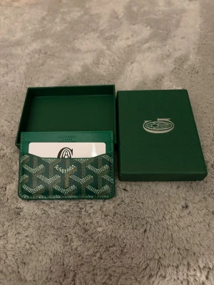 Snygg Grön korthållare  - Snygg grön korthållare med klassiskt mönster i vitt och grönt . Tillverkad i exklusivt läder med flera fack för kort. Kommer med original ask och lock, perfekt för dig som gillar stilrena accessoarer.