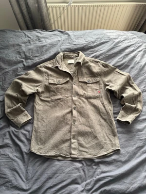 Militärgrön overshirt - Militärgrön overshirt från Royal Denim Division i mjukt, strukturerat tyg. Två bröstfickor med lock och knappar, perfekt lager-på-lager-plagg för en avslappnad look. Modellen på bilden är 197 cm lång och väger ca 81 kg