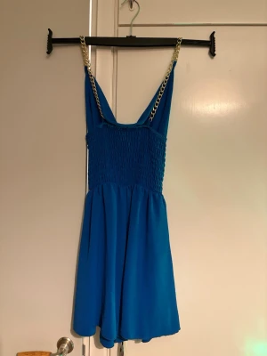 Blå playsuit - Säljer en blå playsuit med tunna axelband i guldfärgad kedja. En snygg och enkel design som passar perfekt till sommaren. Materialet känns lätt och bekvämt.
