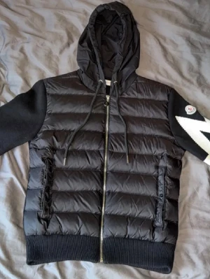 Svart Moncler pufferjacka med huva - Säljer en svart pufferjacka från Moncler med huva och dragkedja. Jackan har stickade svarta ärmar med en vit blixtdetalj på ena ärmen och Moncler-logga på axeln. Kroppen är quiltad och vadderad, perfekt för kalla dagar. Två fickor framtill med knappstängning.
