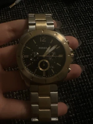 Fossil klocka i guld och silver - Stilren Fossil klocka med svart urtavla och detaljer i guld. Armbandet är i tvåtonad metall, både guld och silver, och har Fossil-loggan på spännet. Klockan har flera små urtavlor och markerade index.