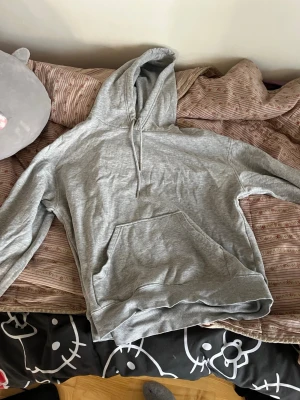 Grå basic hoodie med huva - En enkel och stilren grå hoodie med huva och dragsko. Hoodien har en stor magficka framtill och ribbade muddar vid ärmslut och nederkant. Perfekt för en avslappnad look och passar till det mesta. Tillverkad i mjukt bomullsmaterial.