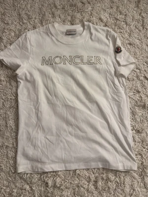 Vit Moncler t-shirt med logga - Snygg vit t-shirt från Moncler med stor logga tryckt över bröstet och Moncler-märke på ärmen. Klassisk passform och rund halsringning, perfekt till jeans eller shorts. Tillverkad i mjuk bomull för skön känsla hela dagen.