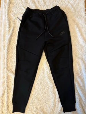 Svarta Nike Tech byxor - Svarta Nike Tech mjukisbyxor med breda muddar vid benslut och midja. Byxorna har snörning i midjan med svart och vit mönstrad dragsko samt fickor på sidorna. Perfekta för en avslappnad stil.