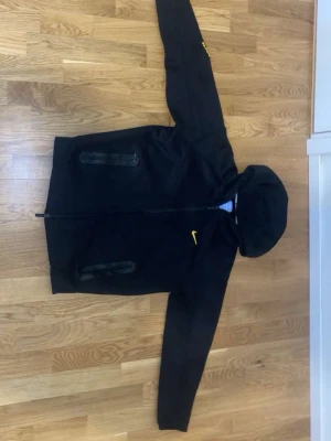 Svart Nike hoodie med dragkedja - Snygg svart hoodie från Nike med dragkedja framtill och två fickor med dragkedja. Har en gul Nike-logga på bröstet och små gula detaljer på ryggen. Tillverkad i mjukt material och har en klassisk huva. Perfekt för en sportig och avslappnad stil.
