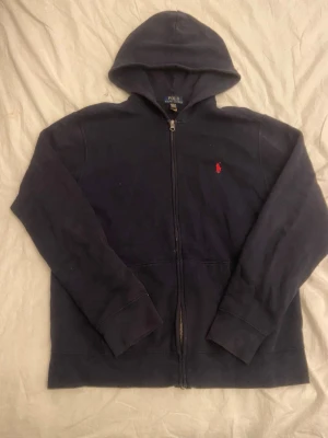 Mörkblå hoodie från Polo Ralph Lauren - Mörkblå hoodie med dragkedja från Polo Ralph Lauren. Klassisk design med huva och det ikoniska röda Polo-märket broderat på bröstet. Tillverkad i mjukt material och har långa ärmar samt ribbade muddar. Perfekt för en avslappnad och stilren look.