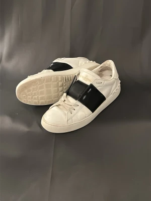 Valentino vita sneakers med svart detalj - Säljer ett par Valentino Open sneakers i vitt skinn med bred svart rand över mitten och gulddetaljer på plösen. Klassisk låg modell med snörning och platt sula. Perfekta för dig som gillar exklusiva och stilrena sneakers.