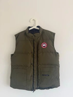 Olivgrön dunväst från Canada Goose - XL - Säljer en olivgrön dunväst från Canada Goose med klassisk logotyp på bröstet. Västen har två stora fickor framtill, hög krage och stängs med dragkedja och knappar. Perfekt för lager-på-lager och håller dig varm under kyliga dagar.