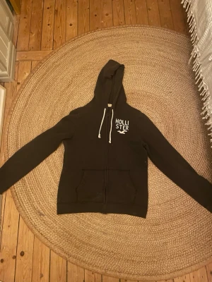 Mörk brun Hollister hoodie med dragkedja - Snygg mörk brun hoodie från Hollister med vit logga och fågel på bröstet. Hoodien har dragkedja framtill, två fickor och vita dragsnören i huvan. Perfekt för en avslappnad och cool stil. Materialet känns mjukt och bekvämt.Är lite fläckig 
