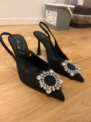 Svarta pumps med strass från Zara - Snygga svarta pumps från Zara med spetsig tå och öppen häl. Skorna har en elegant dekoration framtill med stora glittrande strass-stenar i cirkelform. Klackarna är smala och materialet är i satinliknande tyg.
