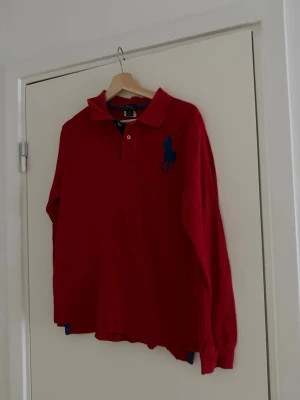 Röd långärmad skjorta från Polo Ralph Lauren - Klassisk röd långärmad skjorta från Polo Ralph Lauren med blå broderad logga på bröstet. Skjortan har krage, knappar vid halsen och är tillverkad i bomull. Perfekt för en stilren och avslappnad look.