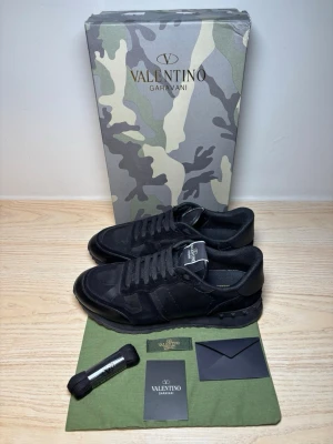 Valentino Rockrunners 43 - Snygga svarta sneakers från Valentino Garavani med diskret camomönster och detaljer i mocka och läder. Klassisk låg modell med svarta skosnören och robust sula. Perfekta för dig som gillar exklusiv streetwear och stilrena detaljer.