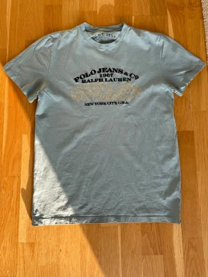 Sällsynt Vintage Polo Jeans Co t-shirt - Snygg ljusblå t-shirt från Polo Jeans Co Ralph Lauren med tryck i svart och beige på bröstet. Klassisk rund hals och korta ärmar. Perfekt för dig som gillar streetwear och vill ha en ikonisk logga på bröstet.