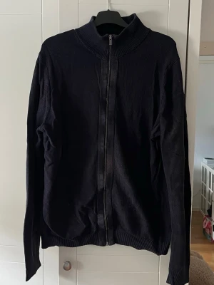 Svart ziptröja med hög krage - En stilren svart ziptröja med hög krage och hel dragkedja framtill. Tröjan har långa ärmar och ribbade muddar vid ärmslut och nederkant. Perfekt för lager-på-lager och enkel att matcha med olika outfits.