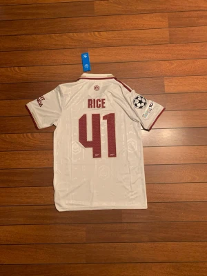 Arsenal bortatröja Rice 41 Adidas - Snygg vit Arsenal bortatröja från Adidas med vinröda detaljer och tryck. Tröjan har spelarnamnet Rice och nummer 41 på ryggen, Champions League-märke på ärmen och klassisk krage. Materialet är lätt och andas, perfekt för fotboll eller träning.