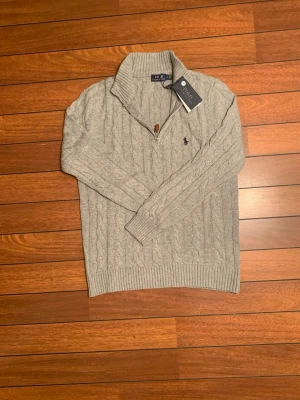 Grå kabelstickad tröja Polo Ralph Lauren - Snygg grå kabelstickad tröja från Polo Ralph Lauren med halv dragkedja och hög krage. Tröjan har det klassiska broderade logot på bröstet och ribbade muddar. Perfekt för lager på lager och en stilren look.