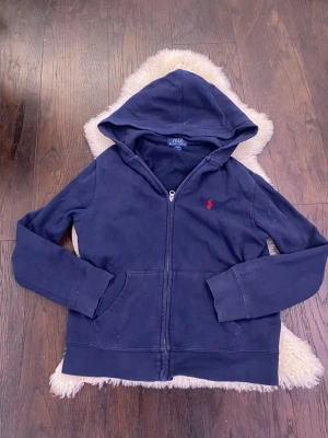 Mörkblå hoodie från Polo Ralph Lauren - Mörkblå hoodie med dragkedja från Polo Ralph Lauren. Klassisk design med huva, kängurufickor och röd broderad logga på bröstet. Hoodien är i storlek L barn så den passar typ om du är 160 elelr kortare skulle jag säga. Mjuk bomullsblandning som är perfekt för chill dagar. Passar dig som gillar stilrena och tidlösa plagg. Kom dm för prisförslag bilder elelr frågor! Säljer billigt som vanligt så passa på!