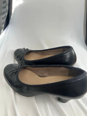 Svarta ballerinaskor med liten klack - Klassiska svarta ballerinaskor i skinn med rund tå och liten klack. Skorna har en söt rosett framtill och beige innersula. Perfekta för dig som gillar en enkel men stilren look.