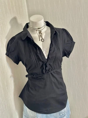 Svart blus med puffärm och knytband - Stilren svart blus med puffiga korta ärmar, krage och v-ringning med volangdetaljer. Blusen har knytband i midjan för en figurnära passform och dekorativa sömmar framtill. Perfekt att matcha med jeans eller kjol för en trendig look.