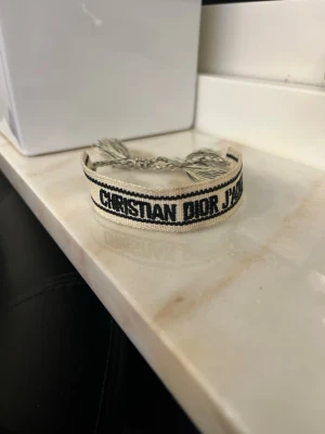 Beige tygarmband från Dior - Snyggt beige tygarmband från Christian Dior med svart broderad text 'Christian Dior J'Adior'. Armbandet har justerbara tofsar i ändarna och är tillverkat i vävt tygmaterial. Perfekt accessoar för att lyfta din outfit. Jättesnyggt armband!