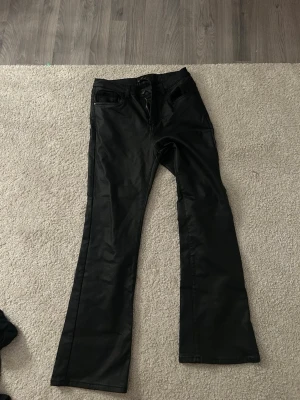 Svarta skinnbyxor byxor  - Snygga svarta byxor från Chilla med bootcut-modell och hög midja. Byxorna har fem fickor, bälteshällor och dragkedja med knapp framtill. Materialet har en lätt glansig finish som ger en cool look.