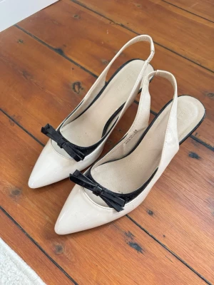 Beiga slingback-pumps med svart rosett - Snygga pumps från Reserved i beige lack med spetsig tå och svart lackdetalj samt rosett framtill. Modellen är slingback med öppen häl och har en smal klack som ger en elegant look. Perfekta för dig som gillar klassisk och stilren design. Lite slitna på klacken. Klacken är ca 7cm💞