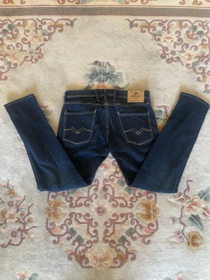 Mörkblå jeans från Replay Anbass - Snygga mörkblå jeans från Replay med raka ben, silverfärgade pipeline sopadlar och en läderpatch. (Midjan 44cm) (Längd 104cm) (Benöppning 17 cm)