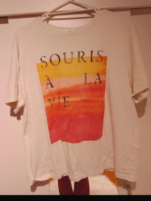 Vit t-shirt med färgglatt tryck - Vit t-shirt med korta ärmar och ett stort färgglatt tryck i gult, orange och rött på framsidan. Texten 'Souris à la vie' är tryckt över motivet. T-shirten har en avslappnad passform och rund halsringning. Perfekt för dig som gillar statement-plagg. Har en liten fläck vid ena ärmen