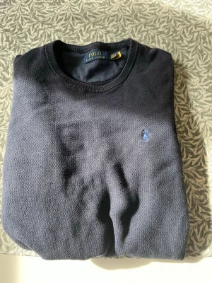 Polo Ralph lauren stickad tröja - Mörkblå rundhalsad tr trickad, klassisk tröja från Polo Ralph Lauren med disk soplog claspad på bröstet. Tröjan har ribbade mud dom ärmslut och nederkant, perfekt för en stilren lat look.