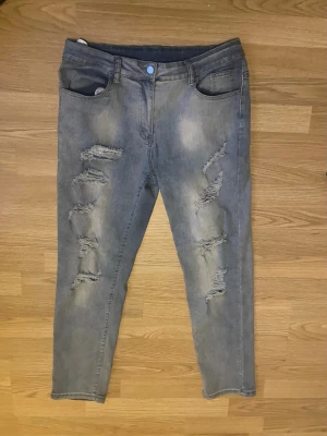 Grå slitna jeans med hål - Säljer ett par grå jeans med flera slitningar och hål på framsidan. Jeansen har klassisk femficksdesign och raka ben. Materialet är denim och färgen är en cool grå nyans. Perfekta för dig som gillar en trendig och avslappnad look.