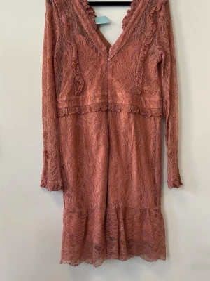 Rosa spetsklänning med volang - Romantisk rosa klänning i spets med v-ringning och volangdetaljer. Klänningen har lång ärm och är tillverkad i ett tunt, genombrutet material som ger en feminin känsla. Perfekt för dig som gillar en söt och trendig stil. Storlek XL men sitter som M. 