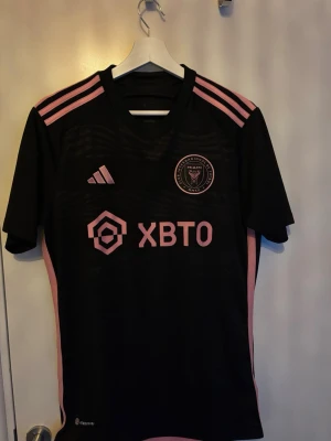 Inter Miami Messi matchtröja svart/rosa - Säljer en svart Inter Miami matchtröja från Adidas med rosa detaljer och logga. Tröjan har Messi och nummer 10 på ryggen, 