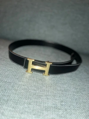 Sv administration skärp med soecifik H sopänne - Sny inf administration skärp med ett ikoniskt H-formad spänne i guld. Bältet är svart och gjort i slätt skinn, perfekt för att ly inf look. Passar till jeans eller hoodies för en trendig vibe.