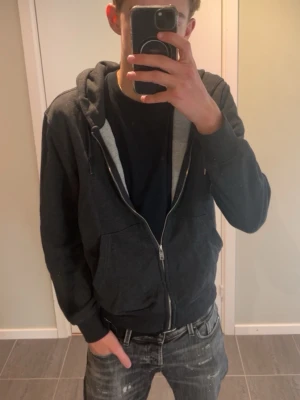 Grå Grish hoodie  - Mörkgrå hoodie med dragkedja och huva. Klassisk modell med fickor framtill och ribbade muddar vid ärmslut och nederkant. Insidan är ljusare grå och materialet känns mjukt och bekvämt, perfekt för chill dagar. Passar till det mesta och är enkel att matcha.