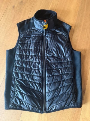 Svart dunväst från Parajumpers - Snygg svart dunväst från Parajumpers med hög krage och gul detalj vid halsen. Västen har dragkedja framtill, två sidofickor och en bröstficka med dragkedja. Tillverkad i lätt, quiltat material som håller värmen utan att kännas klumpig.