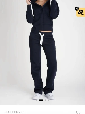 Mörkblå cropped hoodie med dragkedja - Mörkblå cropped hoodie med dragkedja framtill och vita snören i huvan. Hoodien har långa ärmar, ribbade muddar och en avslappnad passform. Tillverkad i mjukt sweatshirtmaterial som är perfekt för en chill och sportig stil.