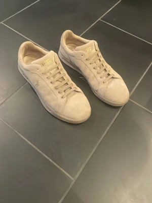 Beige sneakers från Polo Ralph Lauren - Snygga beige sneakers från Polo Ralph Lauren med klassisk låg profil och diskret logga på sidan. Skorna har ovandel i mocka och matchande beige snörning. Perfekta för dig som gillar stilrena och tidlösa sneakers.