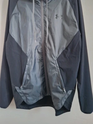 Grå vindjacka från Under Armour - Snygg grå vindjacka från Under Armour med dragkedja framtill, två sidofickor med dragkedja och justerbar huva. Jackan har en sportig design med kontrasterande material och diskret logga på bröstet. Perfekt för blåsiga dagar.