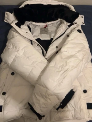 Moncler jacka  - Säljer en vit moncler jacka till ett mycket bra pris, jackan själv håller värmen och är mycket snygg och har en stilren svart moncler symbol på armen . Den har svart päls runt luvan och ger en unik stil som är mycket snygg dm för mer intresse om jackan osv. Den är lite skadad som syns i bilderna.