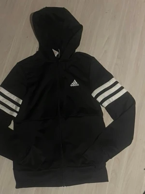 Svart Adidas hoodie med vita ränder - Säljer en svart hoodie från Adidas med dragkedja, huva och vita ränder på ärmarna. Adidas-logga på bröstet och stor logga på ryggen. Perfekt för träning eller chill. Tillverkad i mjukt och stretchigt material.