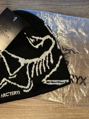Svart mössa från Arc'teryx - Snygg svart mössa från Arc'teryx med vit stickad dinosaurie- och logotypdesign. Tillverkad i mjukt stickat material som sitter skönt på huvudet. Perfekt för dig som vill ha en stilren och sportig look.