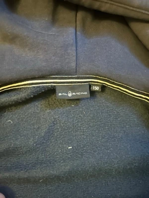 Mörkblå hoodie från Sail Racing - Mörkblå hoodie från Sail Racing med huva och långa ärmar. Tillverkad i mjukt material med fleecefoder på insidan för extra komfort. Klassisk passform och diskreta detaljer. Perfekt för dig som gillar stilrena och funktionella plagg.