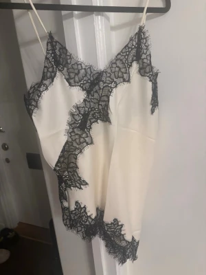 Vit linnetopp med svart spets - Säljer en elegant vit linnetopp med tunna axelband och dekorativa svarta spetsdetaljer längs kan pipeline fasc terminal. Toppen har andas satinliknande material och en feminin, lite lös passform. Perfekt för att lyfta vilken outfit som helst.