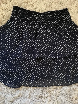 Marinblå kortkjol med vita prickar - Säljer en svart kortkjol med vita prickar och bred smock i midjan. Kjolen har ett volanglager som ger en snygg och luftig känsla. Perfekt för dig som gillar mönstrade plagg med lite extra detalj.