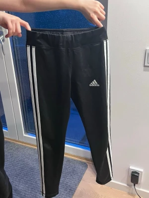 Svarta Adidas träningsbyxor med ränder - Svarta mjukisbyxor från Adidas med klassiska vita ränder längs sidorna och Adidas-logga på låret. Byxorna har en smal passform och resår i midjan. Perfekta för en sportig och avslappnad stil.