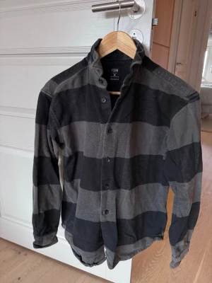 Svart och grå randig flanellskjorta från F5BN - Säljer en flanellskjorta från F5BN med breda svarta och gråa ränder. Skjortan har knappar framtill, långärmad design och en hög krage som ger ett coolt uttryck. 