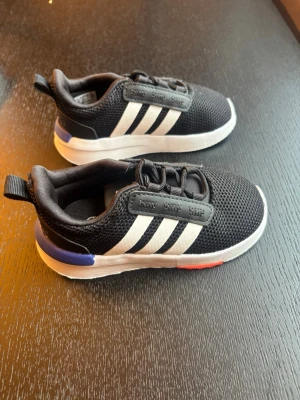 Svarta Adidas sneakers med vita ränder - Snygga svarta Adidas sneakers med klassiska vita ränder på sidorna och detaljer i blått och rött på sulan. Ovandelen är i mesh för bra andningsförmåga och skon har elastiska snören för enkel påtagning. Perfekta för en sportig och avslappnad stil.