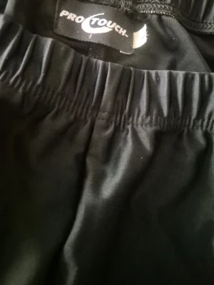 Svarta träningsbyxor Pro Touch - Säljer ett par svarta träningsbyxor från Pro Touch med vita paneler längs sidorna. Byxorna har resår i midjan och är tillverkade i ett stretchigt, snabbtorkande material som passar perfekt för löpning eller gym.